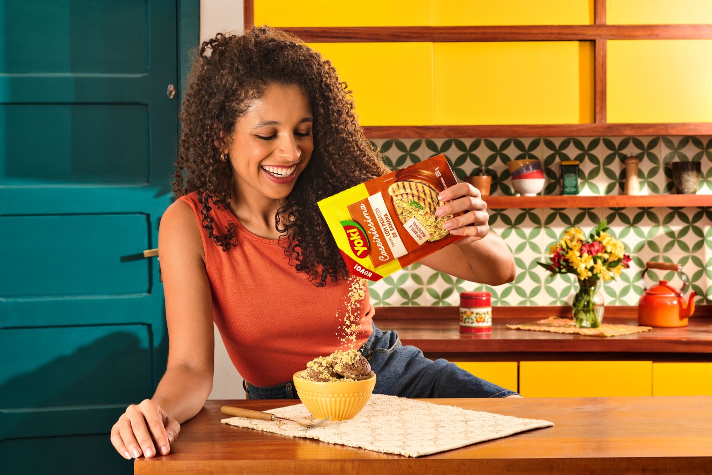 A woman standing in a colorful kitchen pours a packet of instant food into a yellow bowl on a wooden countertop. The kitchen features yellow cabinets, patterned tile backsplash, and various utensils and decorations, including a vase of flowers and a red kettle.
